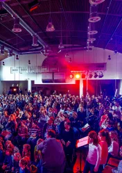 Viele Kinder und Jugendliche sitzen in einer großen, verdunkelten Halle in Reihen und schauen nach vorne. Am Ende des Saals ist ein Scheinwerfer angebracht, der rotes Licht über die Zuschauer strahlt.