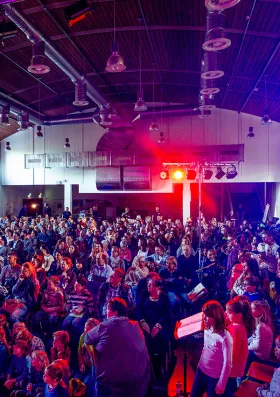 Viele Kinder und Jugendliche sitzen in einer großen, verdunkelten Halle in Reihen und schauen nach vorne. Am Ende des Saals ist ein Scheinwerfer angebracht, der rotes Licht über die Zuschauer strahlt.